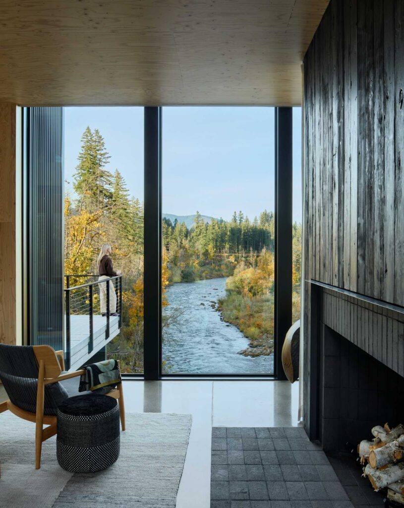 A serene living space framed by floor-to-ceiling windows with views of a river and surrounding forest.