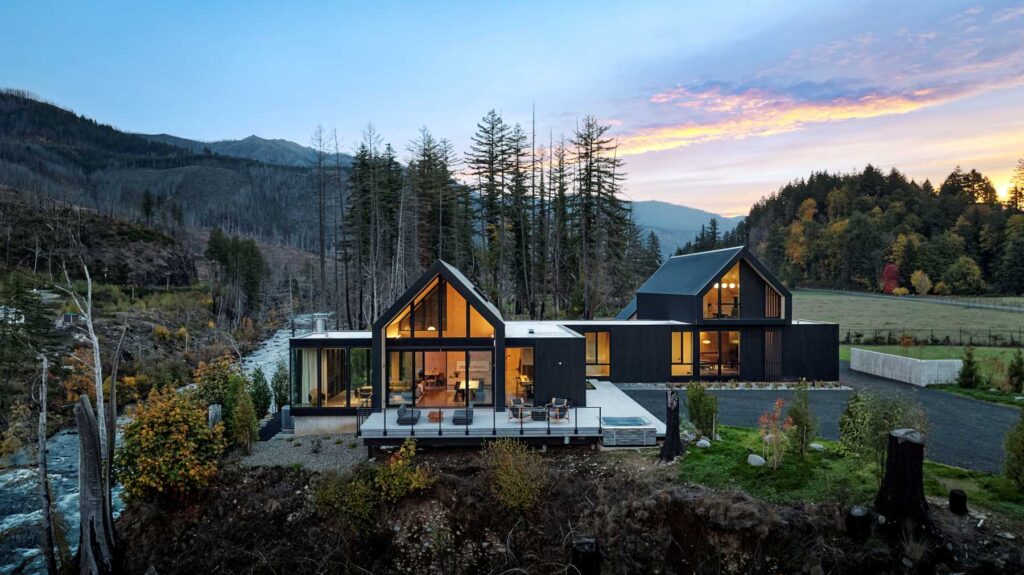 Aerial view of modern homes overlooking a river at sunset, framed by forested hills and mountains.