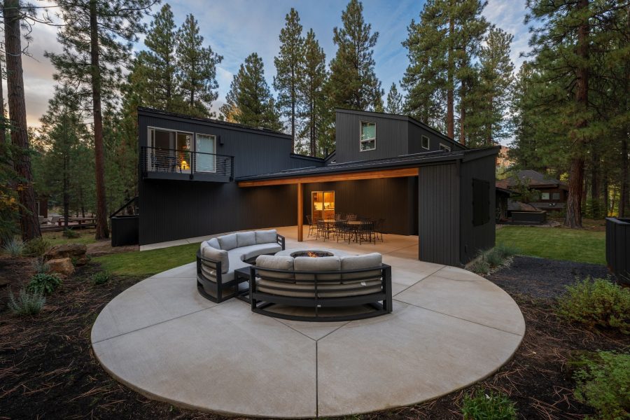 Modern black exterior home in Sisters, Oregon with angular rooflines, covered patio dining area, and circular concrete fire pit lounge surrounded by pine trees.