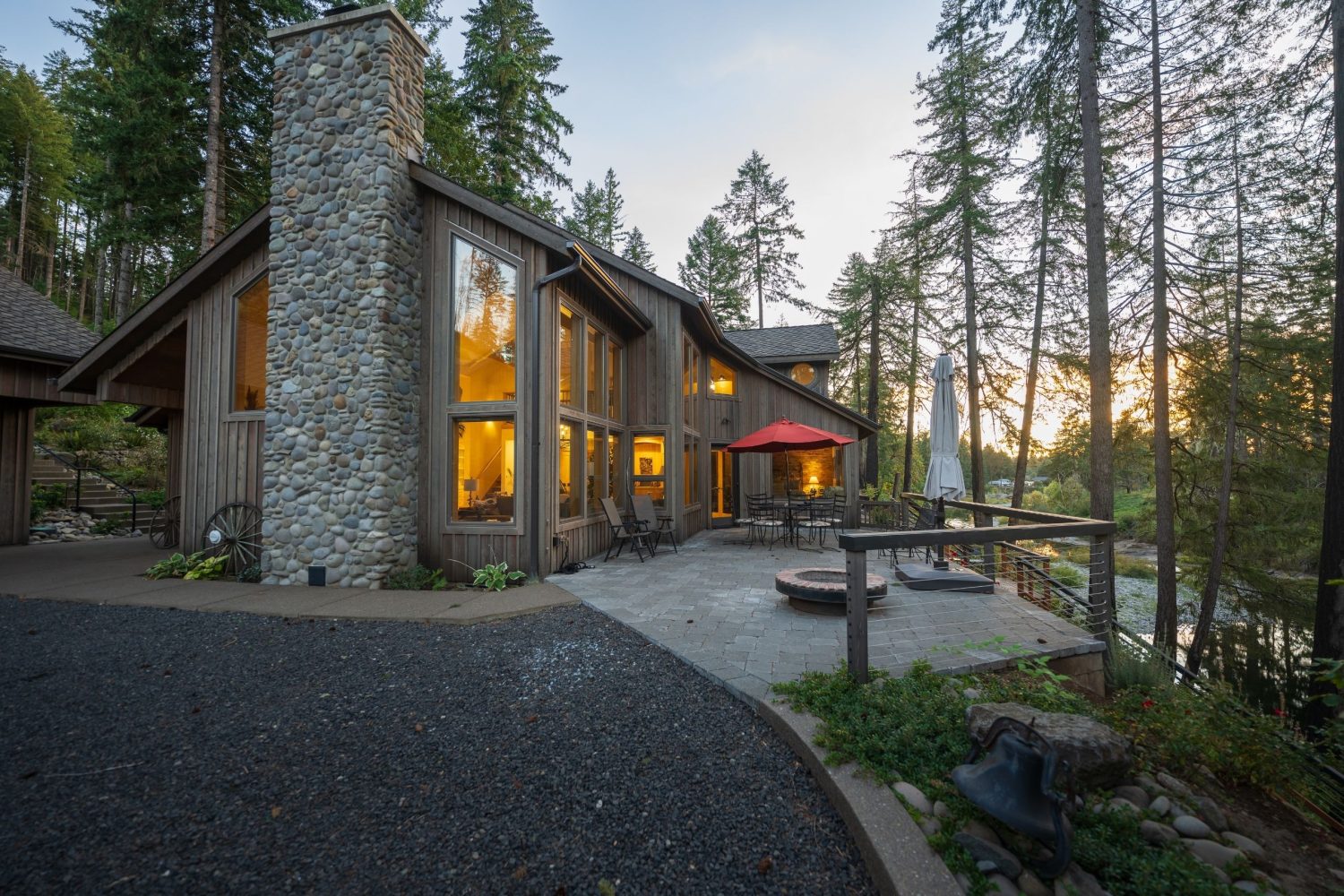 Modern rustic home with stone chimney, large windows, and outdoor patio overlooking a river in Central Oregon.
