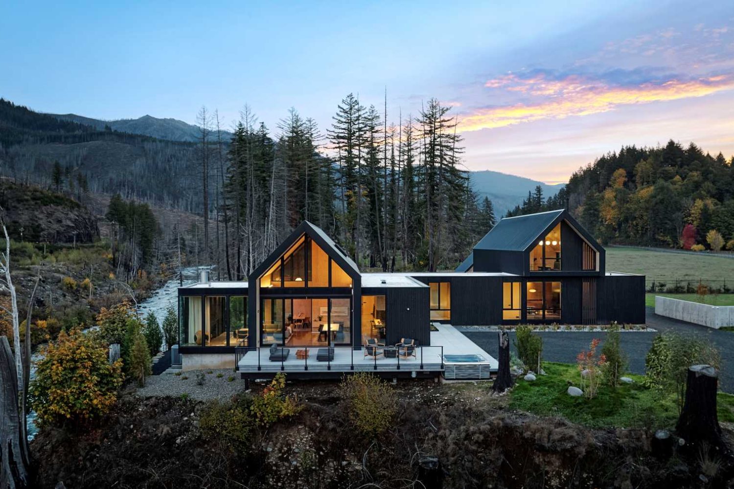 Aerial view of modern black-clad homes overlooking a river at sunset in a forested mountain setting